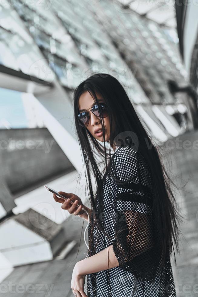Feeling confident. Beautiful young woman in casual wear holding a smart phone while standing outdoors photo