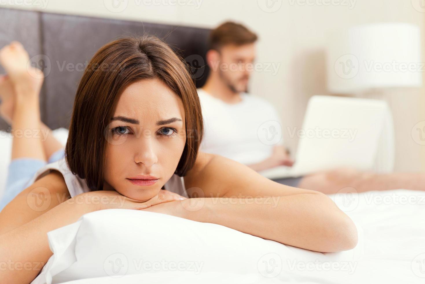 If he only knew. Displeased young woman lying on bed and leaning her face on the pillow while young man working on laptop in the background photo