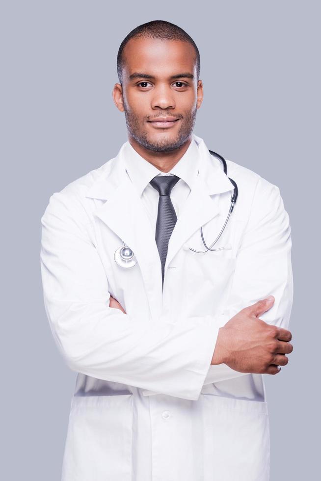 médico varón confiado. médico africano confiado mirando la cámara y manteniendo los brazos cruzados mientras está de pie contra el fondo gris foto
