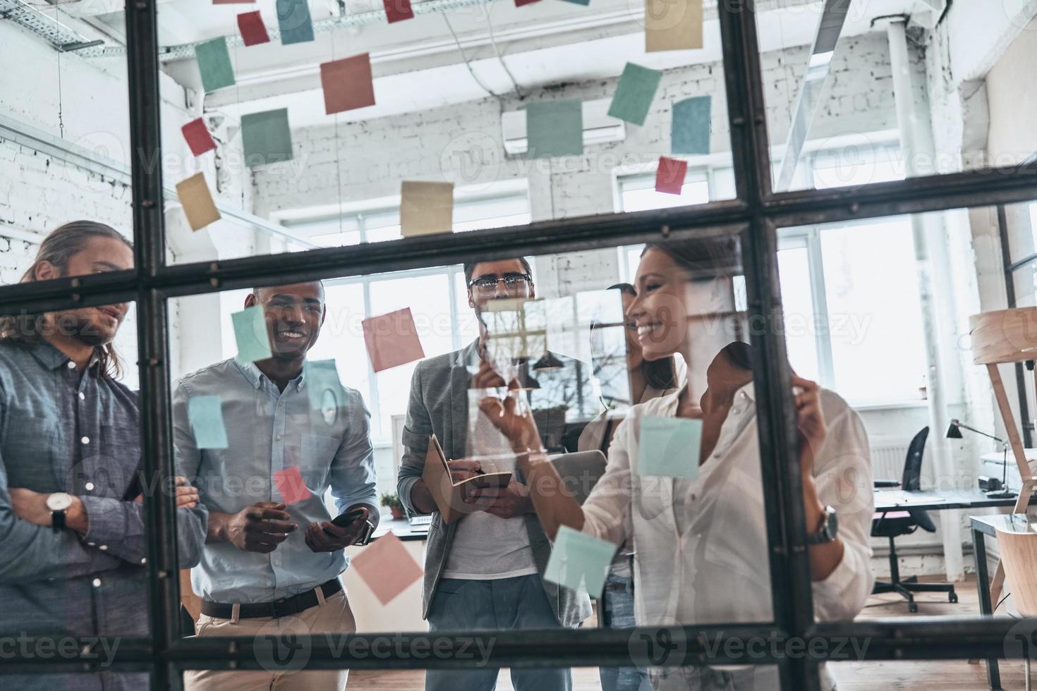 Discussing strategy. Group of young modern people in smart casual wear using adhesive notes while standing behind the glass wall in the board room photo