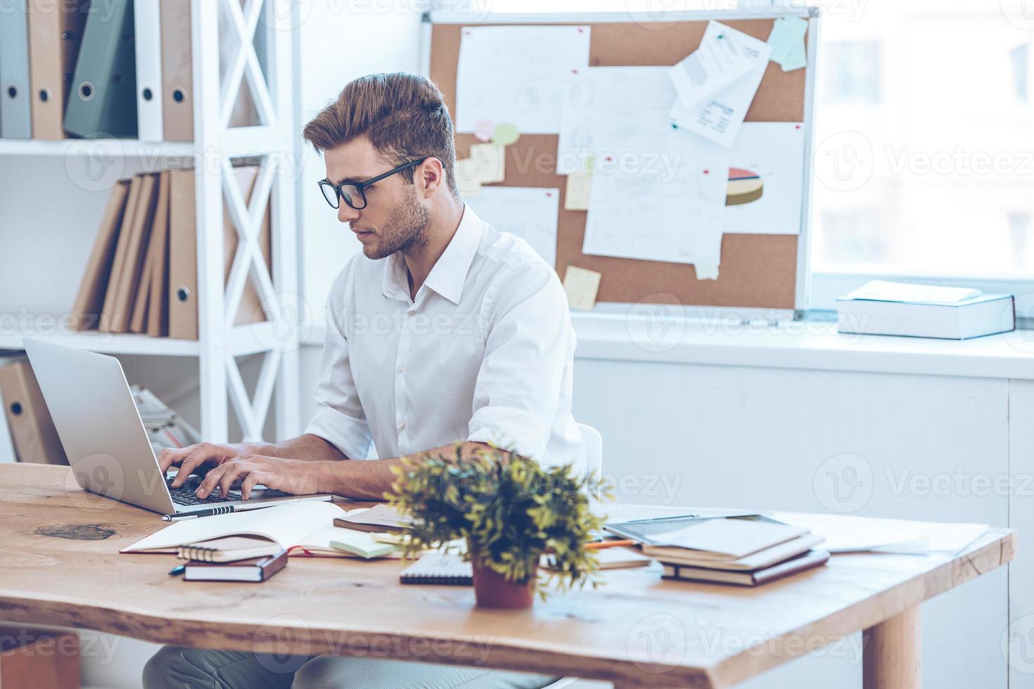 Sending email to his coworkers. Side view of handsome young man in glasses working with laptop while sitting at his working place photo