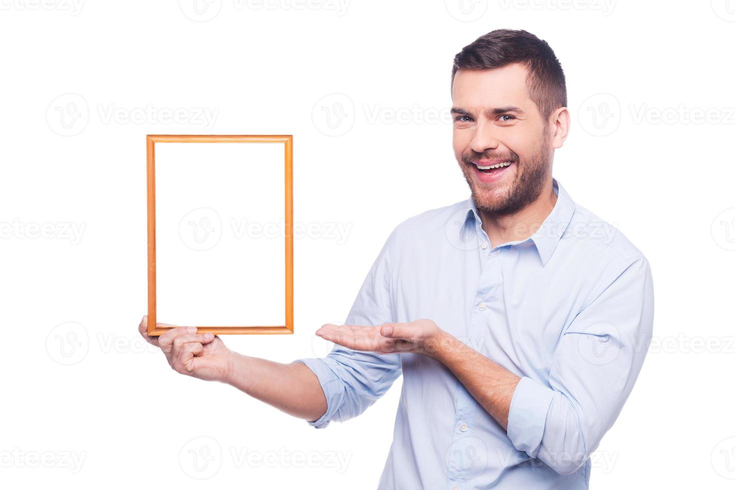 copie el espacio en el marco de la imagen. un joven guapo con camisa sosteniendo un marco de fotos y apuntándolo con una sonrisa mientras se enfrenta a un fondo blanco