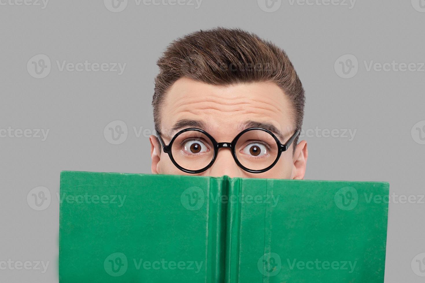 Reading an exciting book. Surprised young man in glasses looking out of the book while isolated on grey background photo