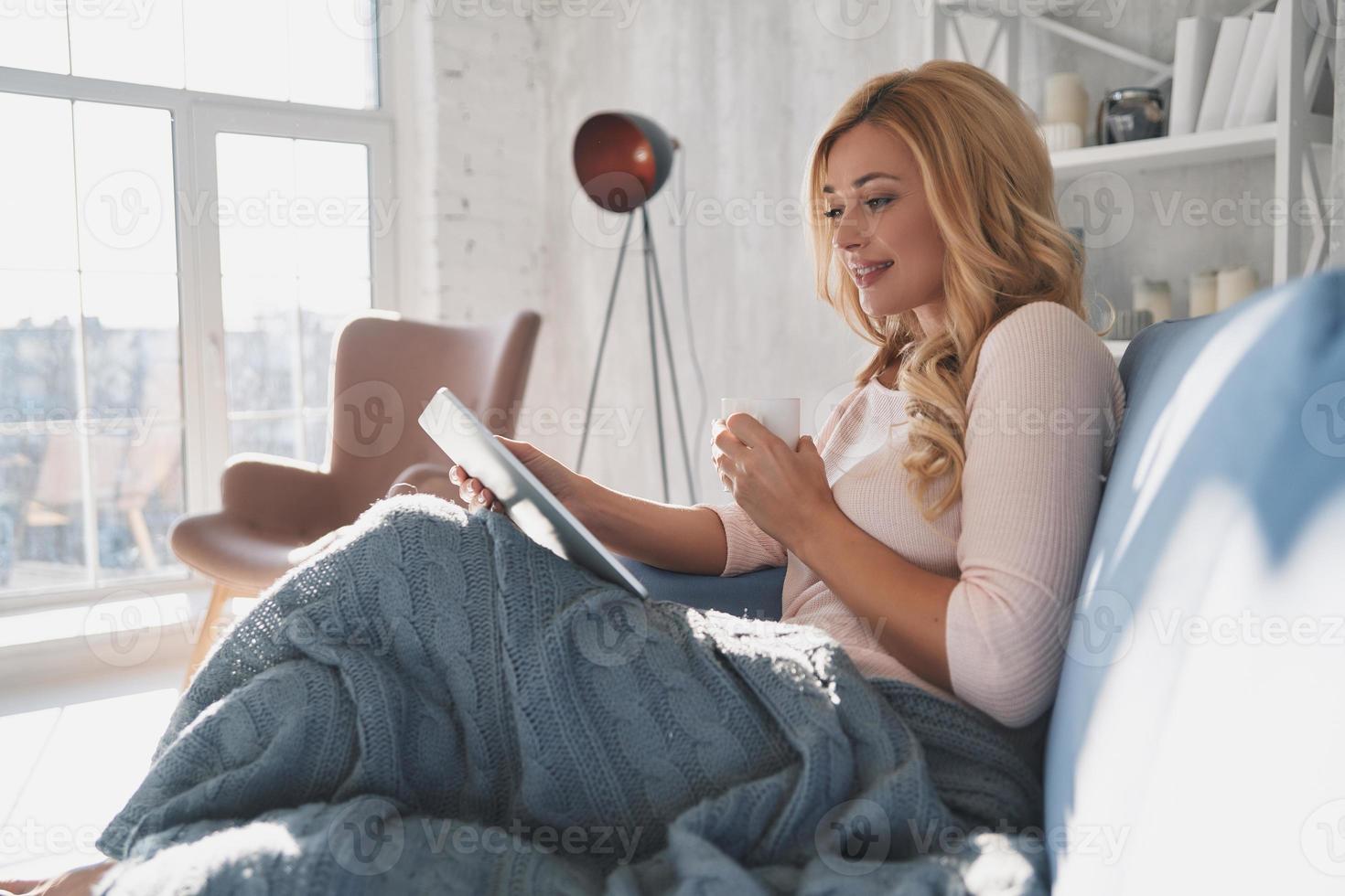 Interesting... Attractive young woman using digital tablet and smiling while sitting on the sofa at home photo