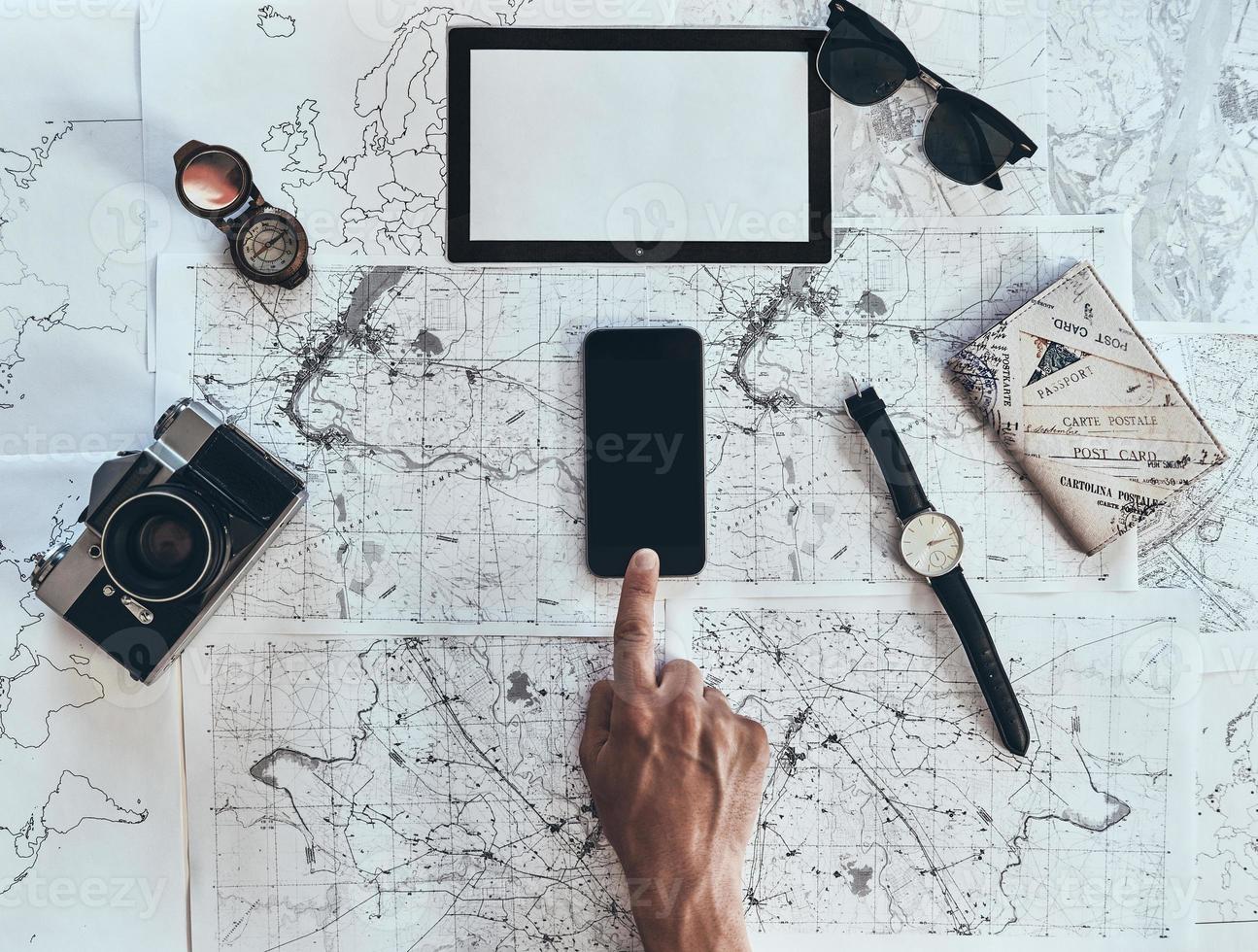 Exploring the world. Close up top view of man using smart phone with sunglasses, photo camera, compass, watch and passport lying on map around