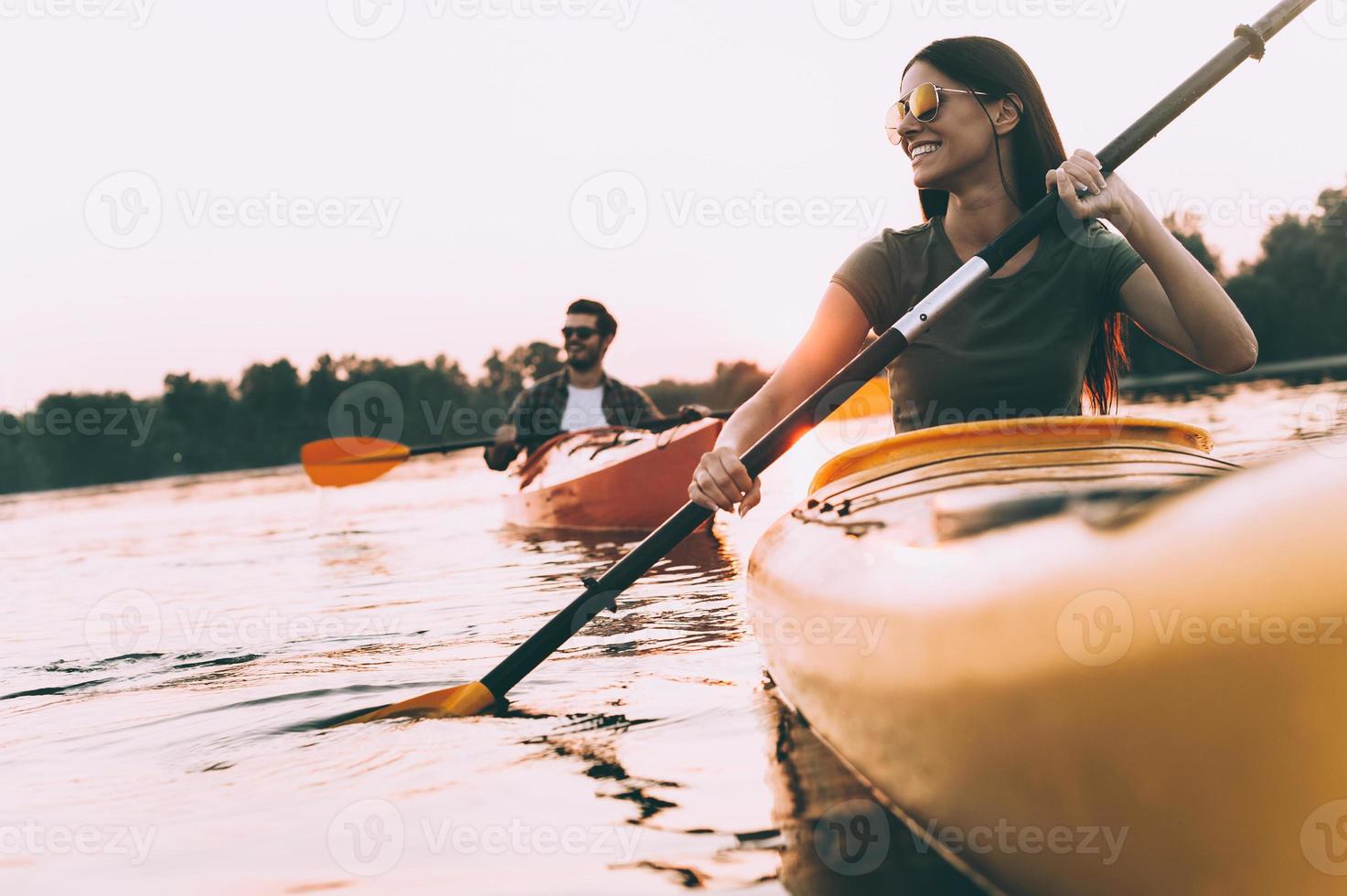 Enjoying nice time on river together. Low angle view of beautiful young couple kayaking on river together and smiling with sunset in the background photo
