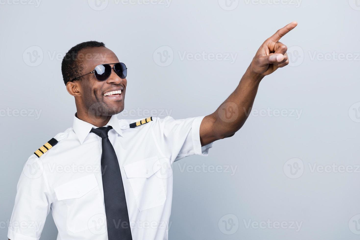 enamorado del cielo. alegre piloto africano uniformado señalando hacia otro lado y sonriendo mientras se enfrenta a un fondo gris foto