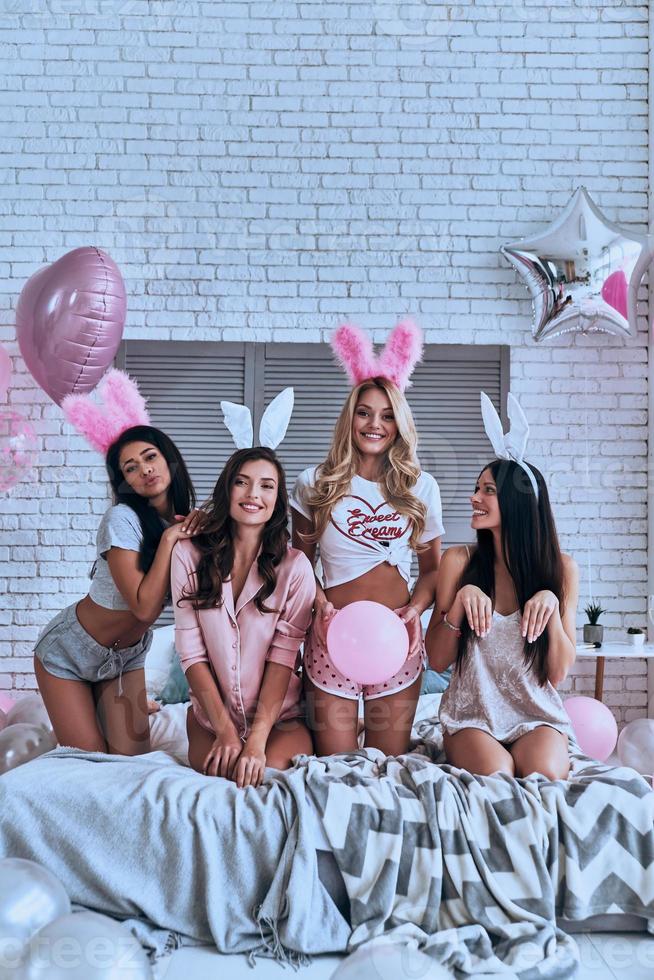 Having fun. Four playful young smiling women in bunny ears making a face and gesturing while sitting on the bed with balloons around photo