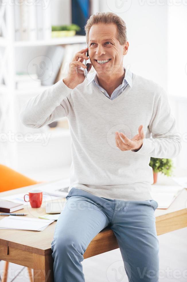 Good business talk. Cheerful mature man talking on the mobile phone and smiling while leaning at the desk in office photo