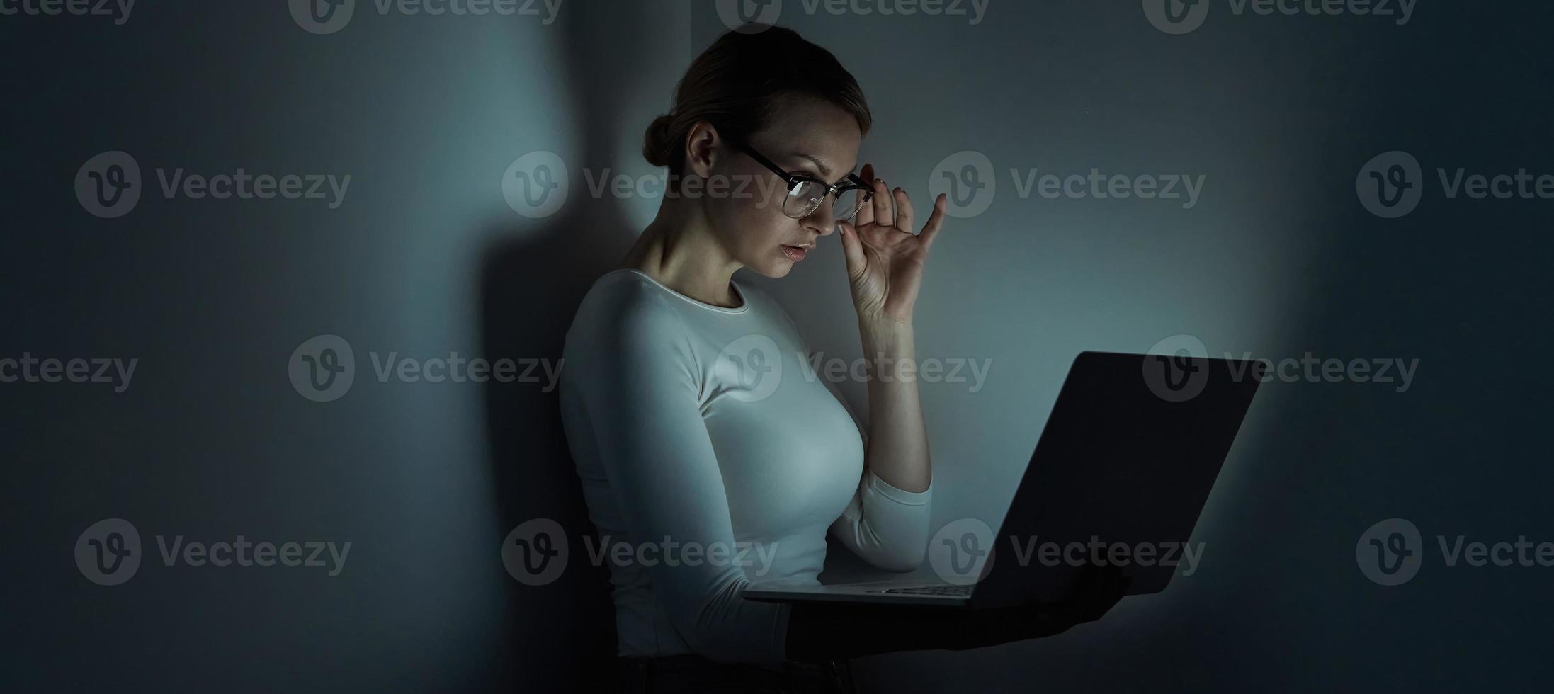 Beautiful young woman adjusting her glasses while working on laptop against a dark wall photo