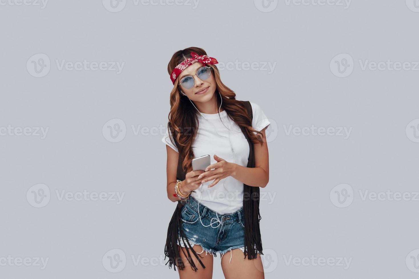 Attractive stylish young woman listening music and smiling while standing against grey background photo