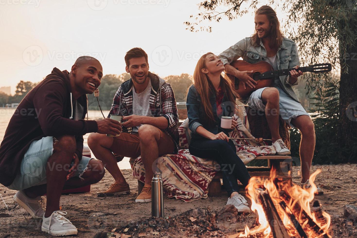 Carefree camping. Group of young people in casual wear smiling while enjoying beach party near the campfire photo