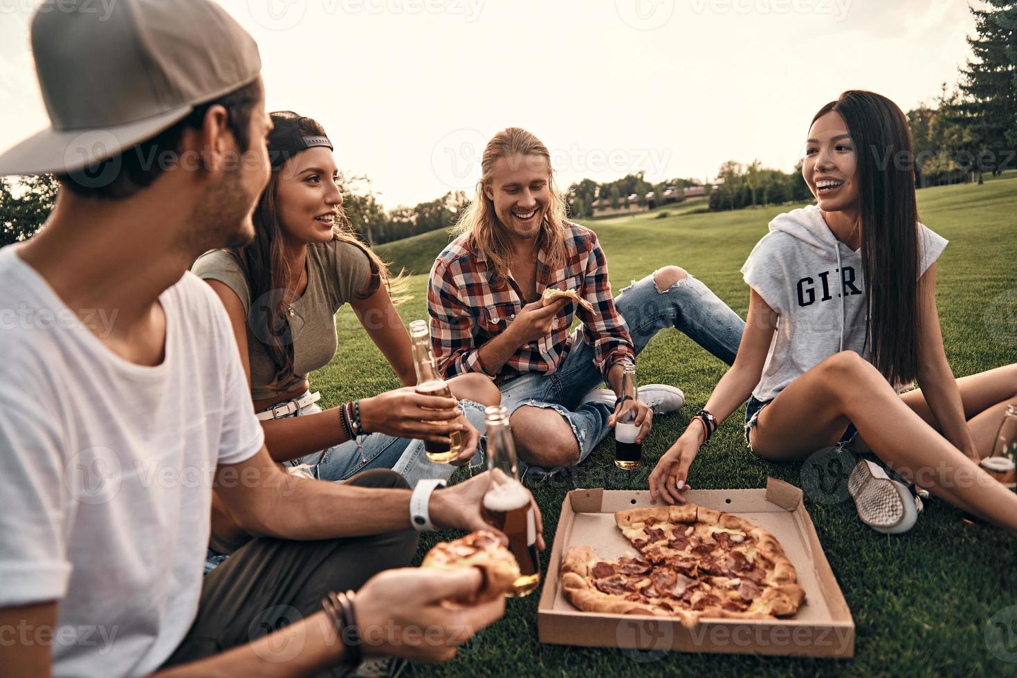Fun time with friends. Group of young smiling people in casual wear enjoying pizza and beer while sitting on the grass outdoors photo