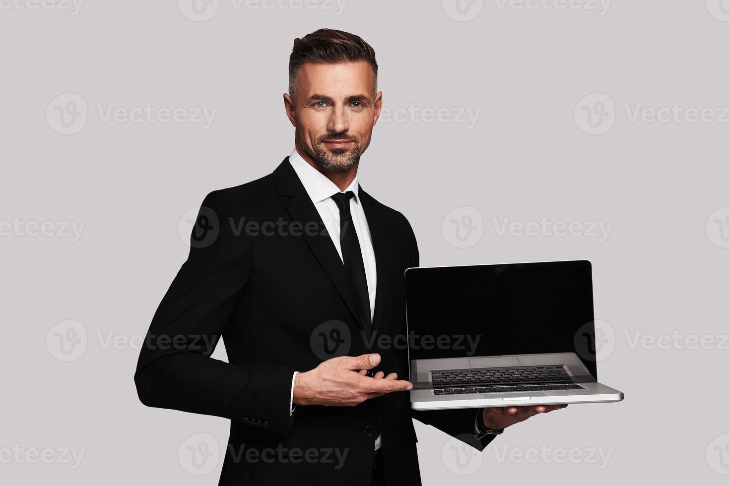 Look over here Handsome young man in full suit pointing copy space on laptop and smiling while standing against grey background photo
