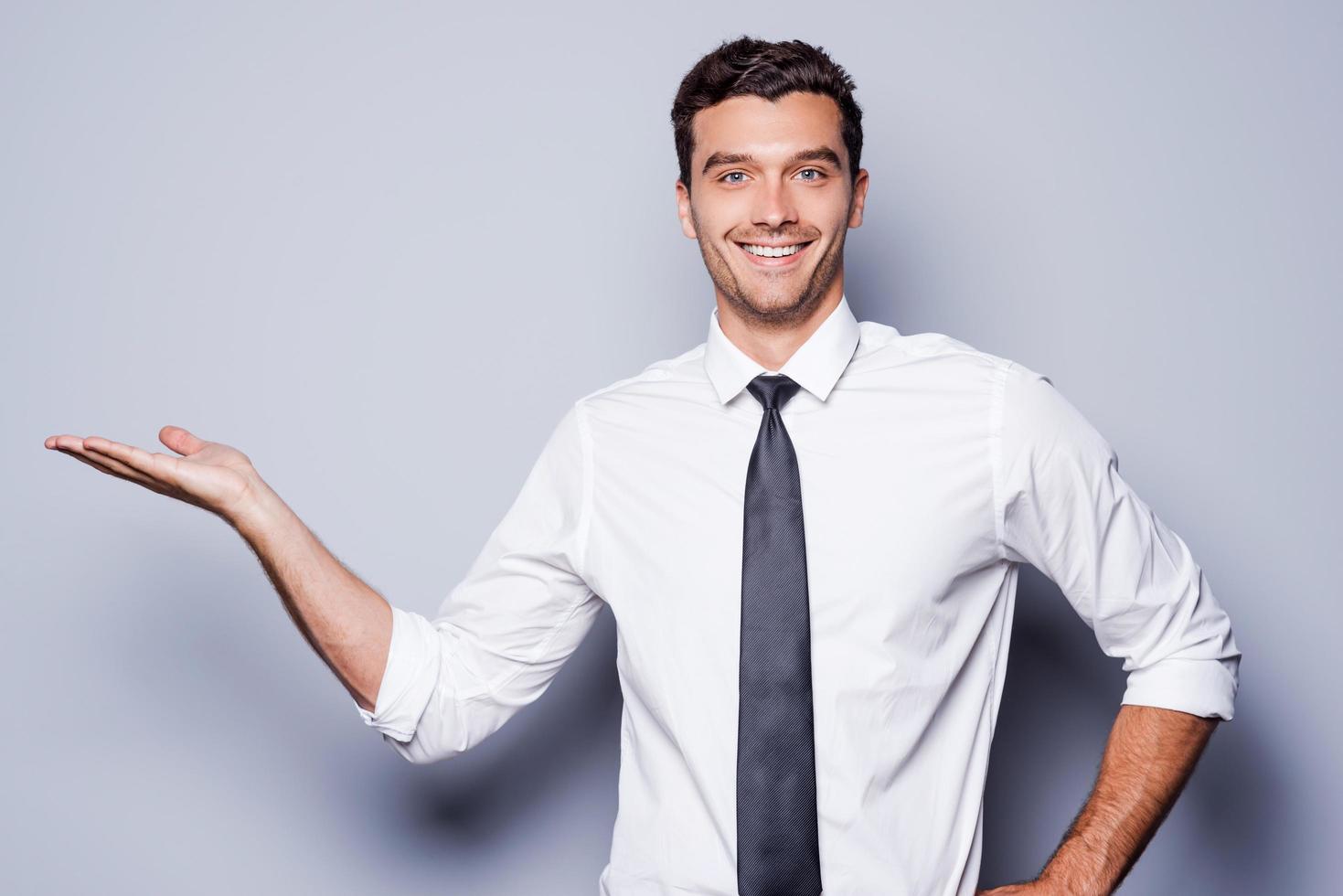 Copy space on his hand. Handsome young man in shirt and tie holding copy space on his hand and smiling while standing against grey background photo
