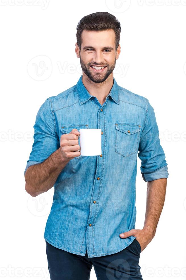 Coffee break. Confident young handsome man in jeans shirt holding cup with hot drink and smiling while standing against white background photo