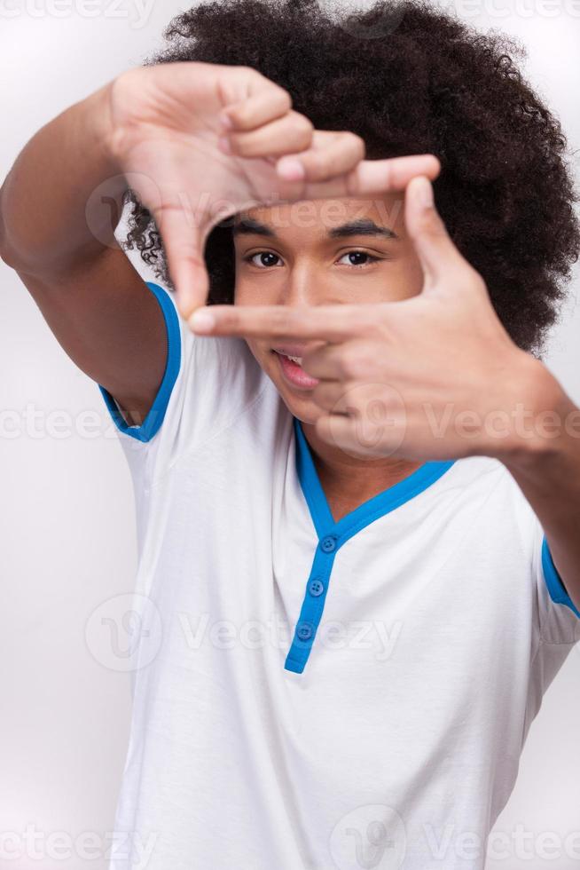 Focusing on you. Cheerful African teenager looking through a finger frame and smiling while standing isolated on grey background photo