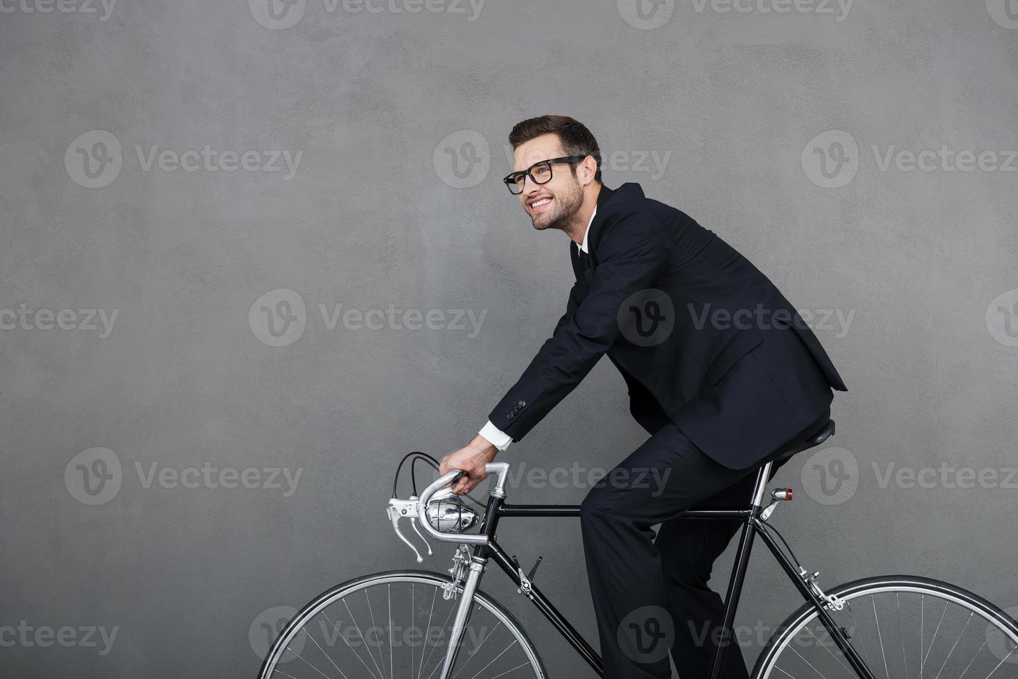 Keep calm and ride on. Cheerful young businessman smiling and looking away while cycling against grey background photo