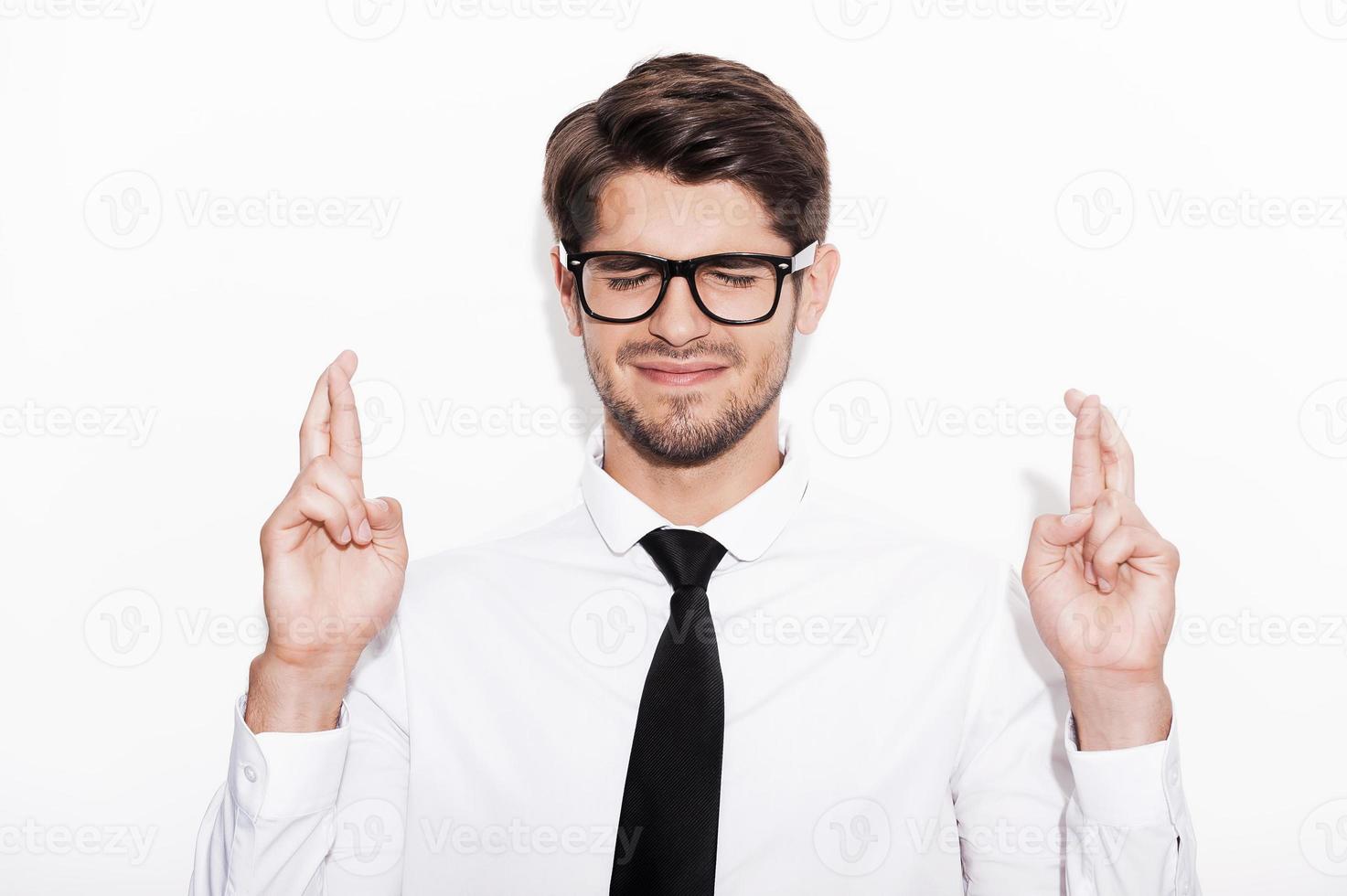 Waiting for special moment. Young man keeping fingers crossed and eyes closed while standing against white background photo