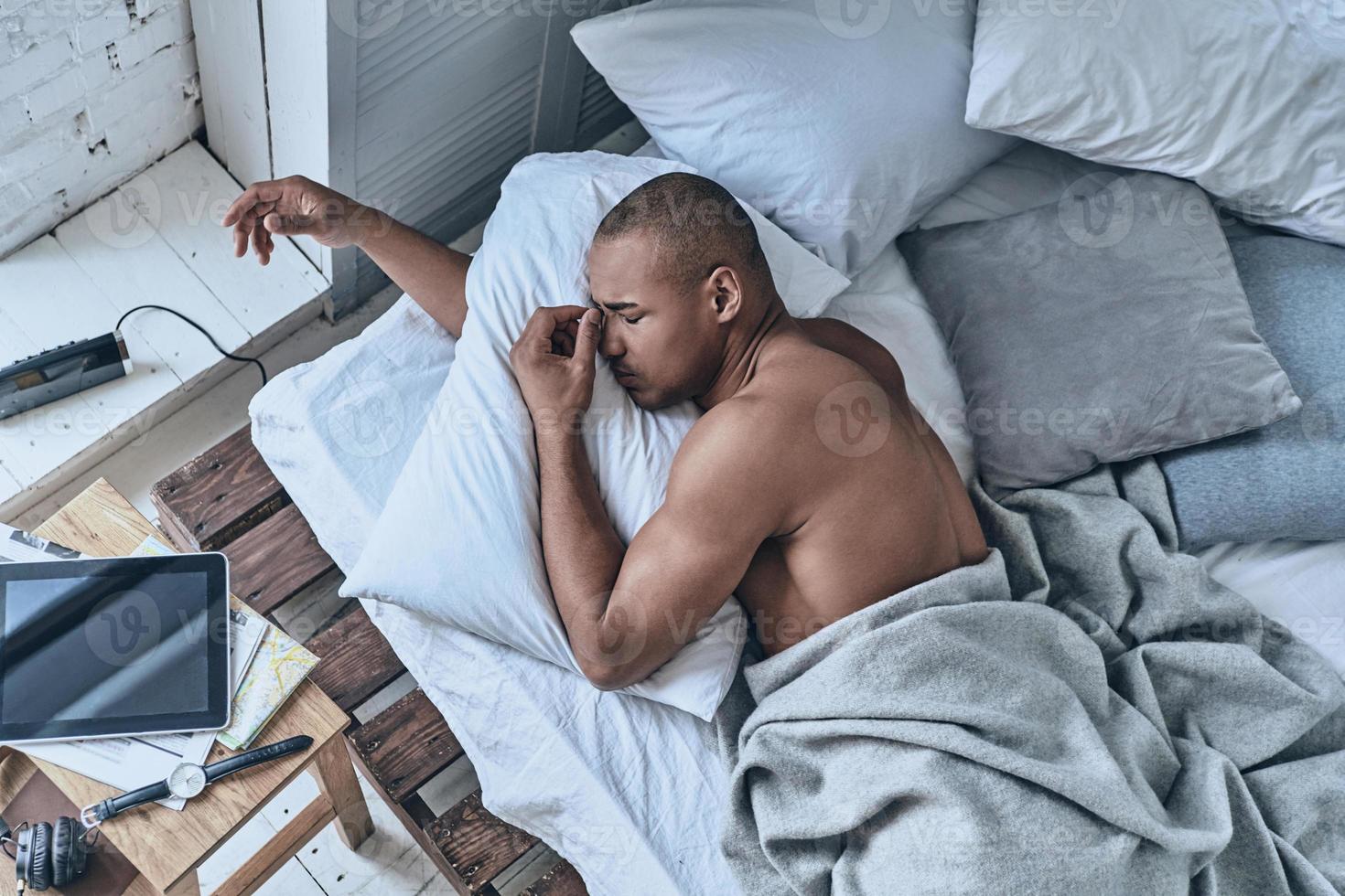 So hard to wake up. Top view of young African man massaging nose while lying in the bed at home photo