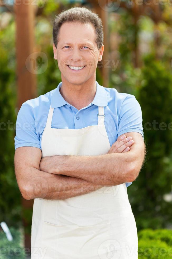 I have the best job in the world Handsome mature man in apron keeping arms crossed and smiling while standing in a greenhouse photo