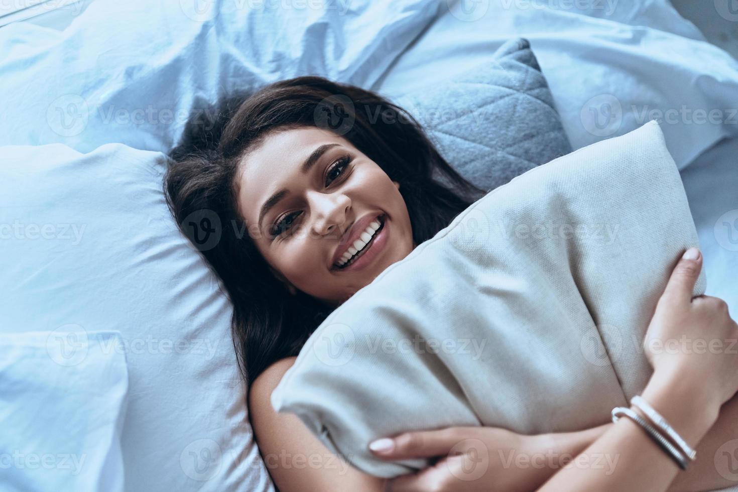 Feeling rested. Top view of attractive young woman embracing pillow and smiling while lying on the bed at home photo