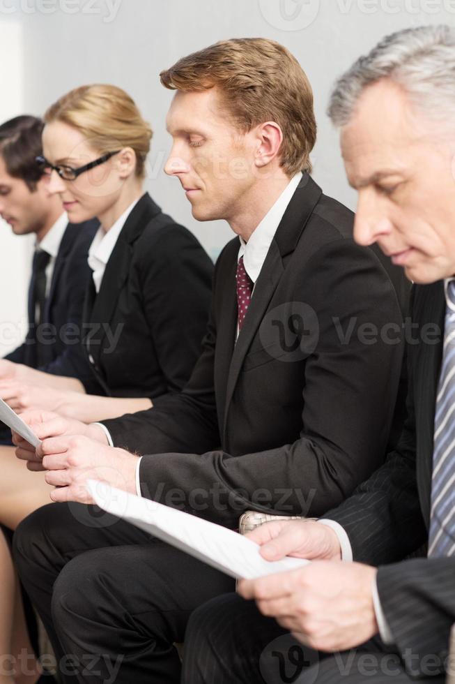 Waiting in line. Four people in formalwear waiting in line while sitting in a row and holding papers in their hands photo