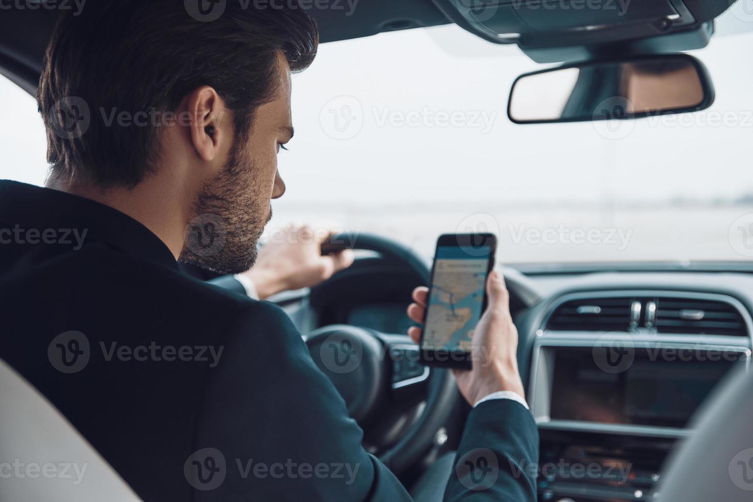 Where am I Rear view of young man in full suit using smart phone to check the map while driving a car photo