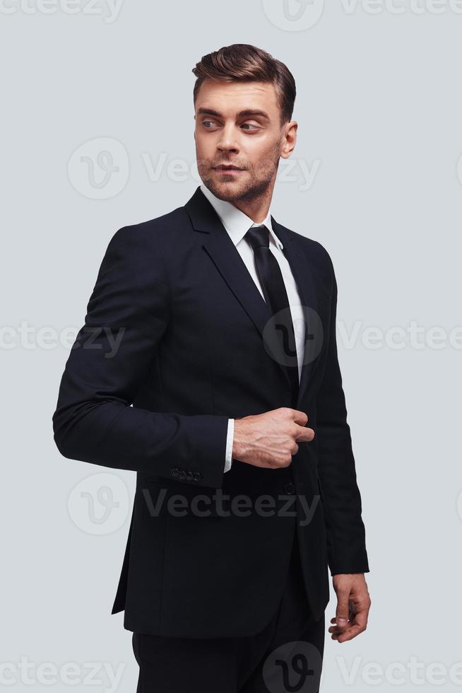 Always perfect. Handsome young man in full suit adjusting his jacket and looking away while standing against grey background photo