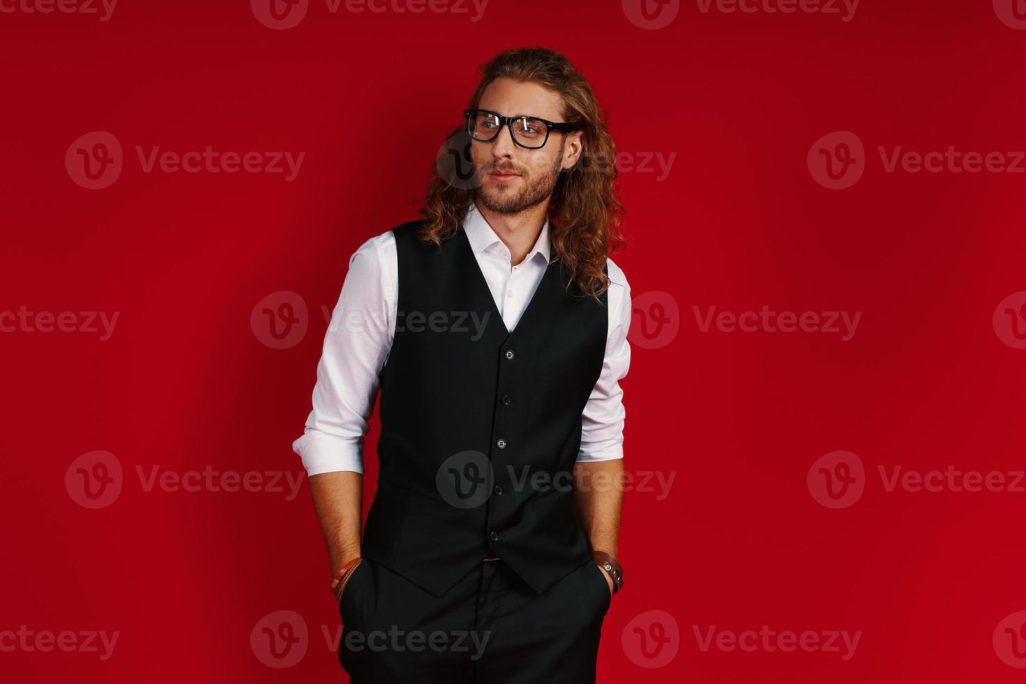 Elegant young man in suit and eyewear looking away with smile while standing against red background photo