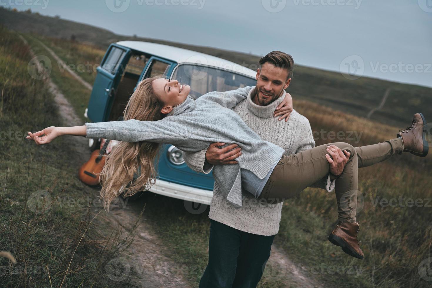 la vida es increíble, un joven apuesto que lleva a su atractiva novia y sonríe mientras está de pie cerca de la mini furgoneta azul de estilo retro foto