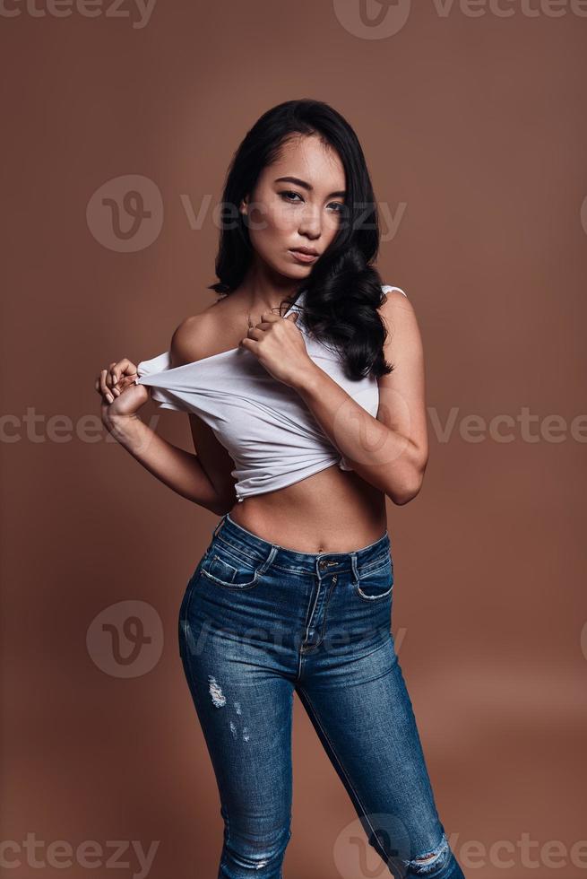 Feeling flirty. Beautiful young woman adjusting her shirt and looking at camera while standing against brown background photo