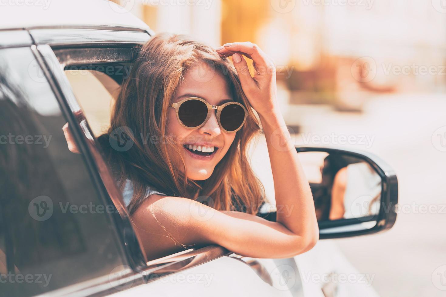 Are you going with me Rear view of beautiful young cheerful women looking at camera with smile while sitting in her car photo