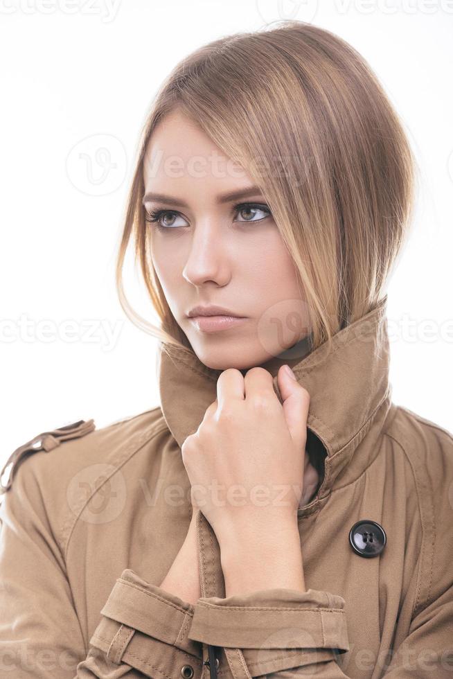 Feeling warm and trendy. Attractive young woman in coat covering her neck with collar and looking away while standing against white background photo