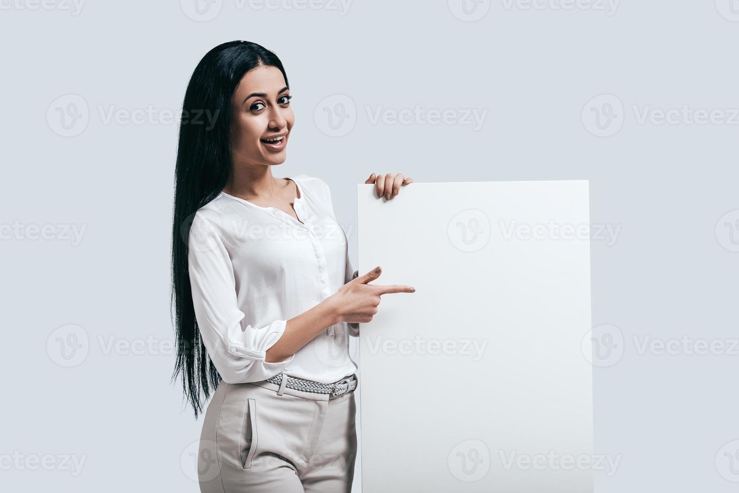 Place new ideas here Young confident woman in white shirt pointing on blank flipchart while standing against grey background photo