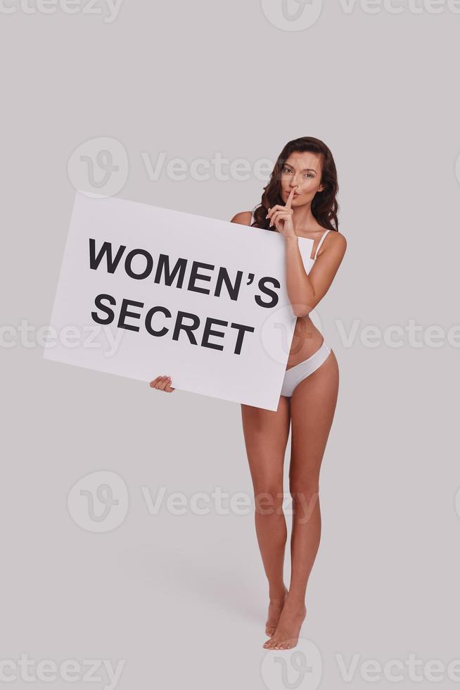 Do not tell anyone.  Full length of attractive young woman holding a poster and keeping index finger by her lips while standing against grey background photo