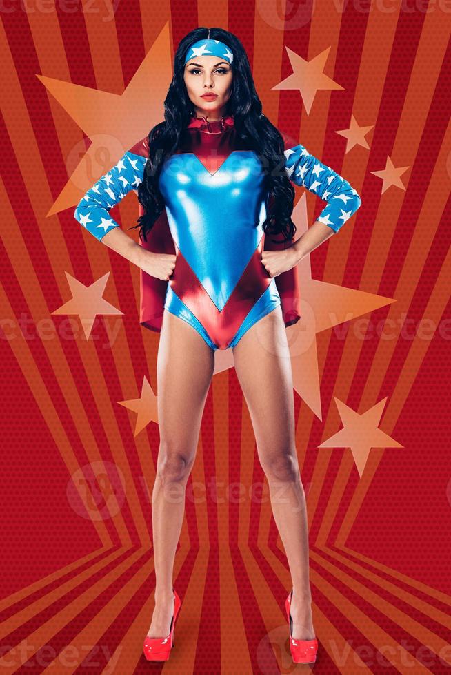 She can fight crime anywhere. Full length of beautiful young woman in superhero costume looking at camera and keeping hands on hips while standing against red background photo