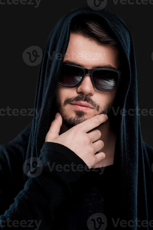 He got dark side. Confident young man in black hood holding hand on chin and looking at camera while standing against black background photo