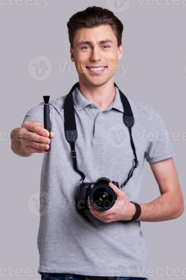 cuidar de su lente joven alegre en camisa de polo sosteniendo una cámara digital y estirando el cepillo de la lente mientras está de pie en el estudio foto