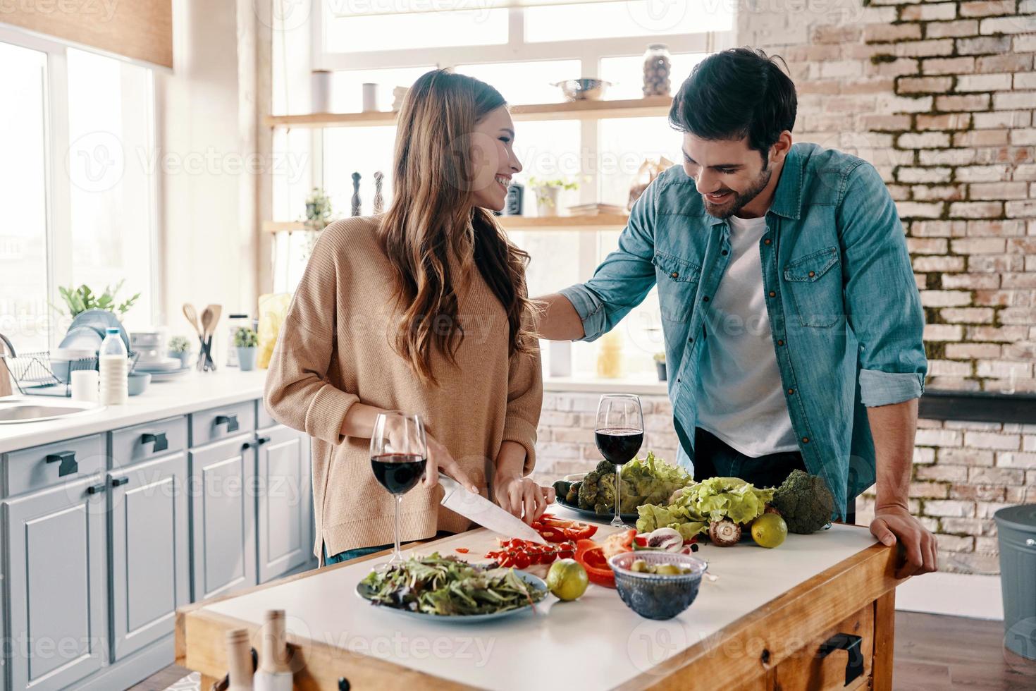 Doing everything together. Beautiful young couple cooking dinner and drinking wine while standing in the kitchen at home photo