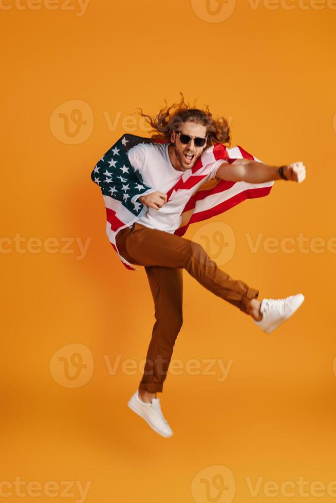 Confident young man in casual clothing making a face and punching while hovering covered in American flag against yellow background photo