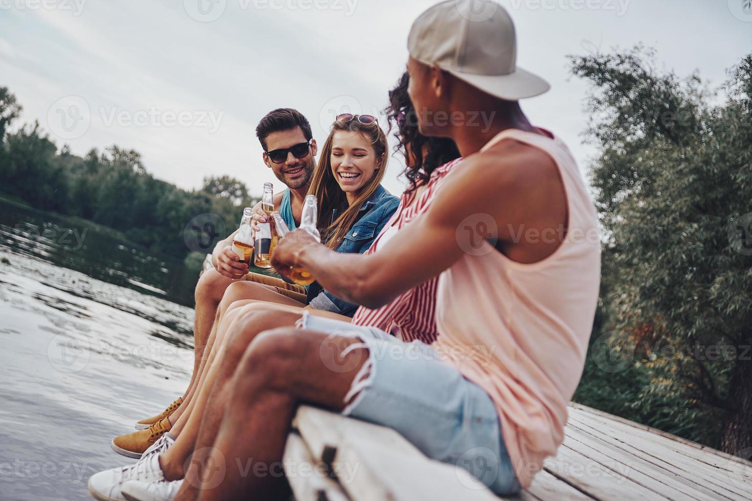 Enjoying every minute together. Group of happy young people in casual wear smiling and drinking beer while sitting on the pier photo