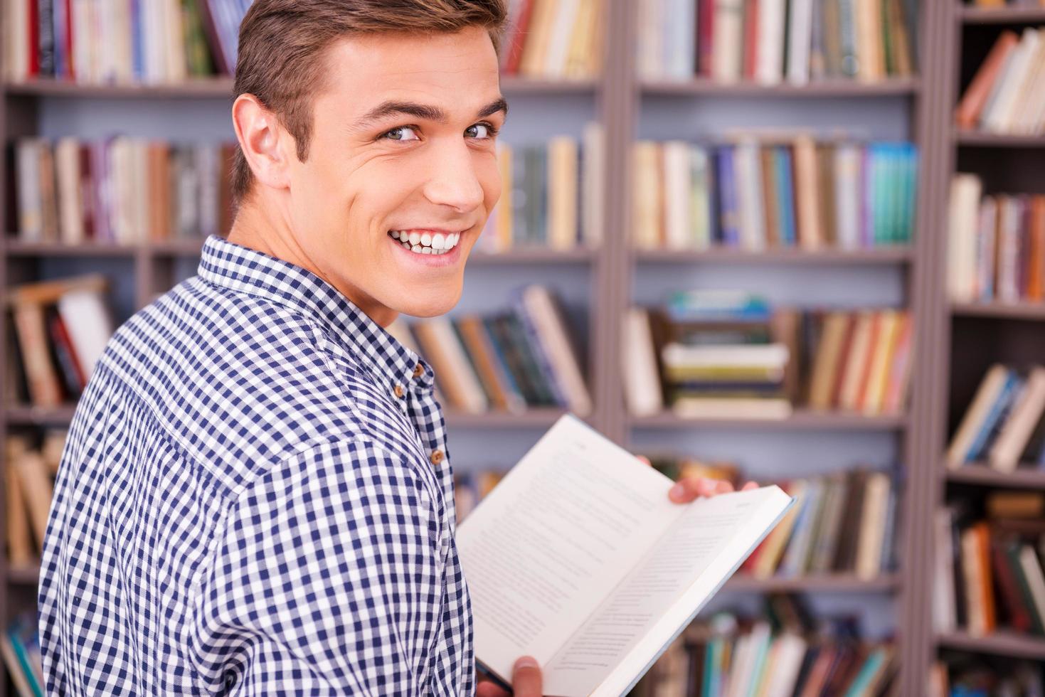 ratón de biblioteca feliz. vista trasera de un joven feliz sosteniendo un libro y mirando por encima del hombro mientras está de pie contra la estantería foto