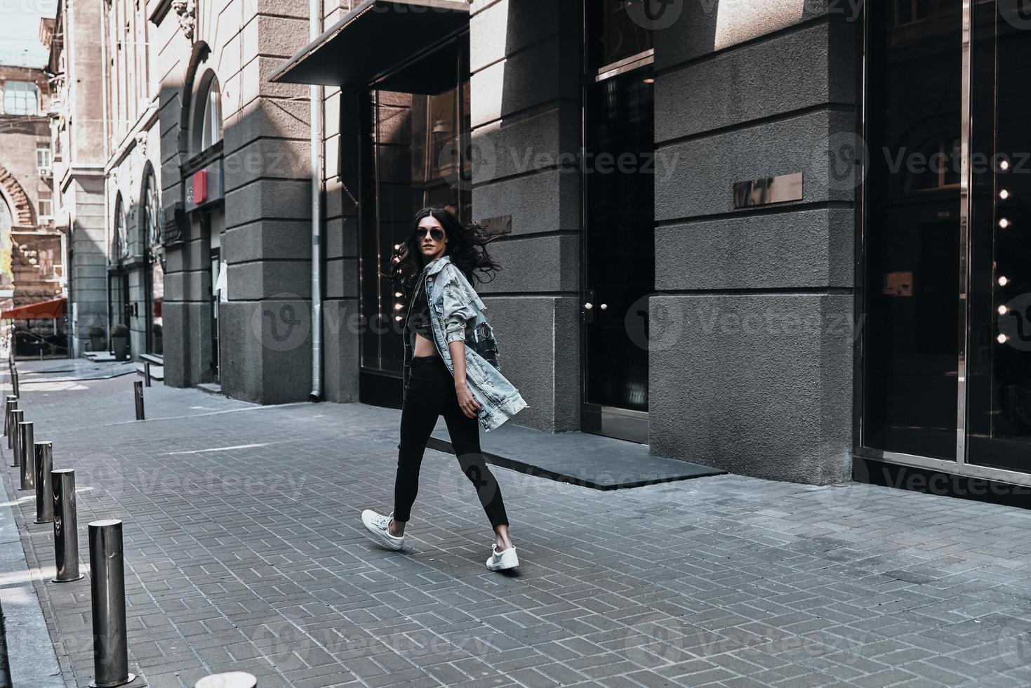 City girl. Full length rear view of young woman looking over her shoulder while walking down the street photo