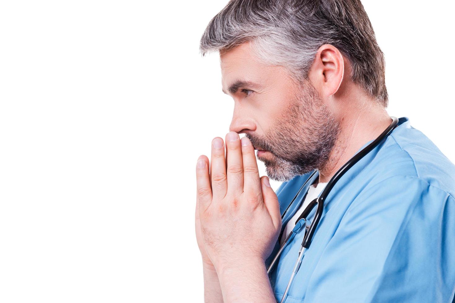 médico rezando. Vista lateral de un médico de pelo gris maduro con las manos juntas cerca de la cara mientras está de pie aislado en blanco foto