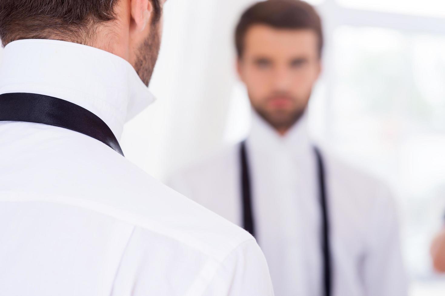 Getting ready for a special day. Rear View of young man in white shirt and untied necktie standing against mirror photo