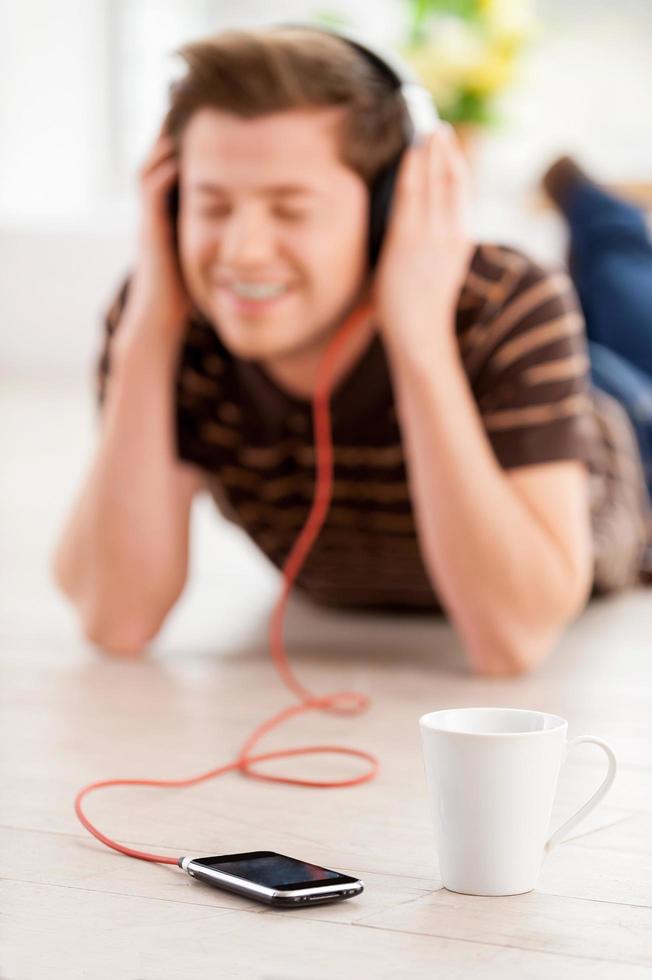 Enjoying his favorite music. Happy young man in headphones listening to the music and keeping eyes closed while lying on the floor at his apartment photo
