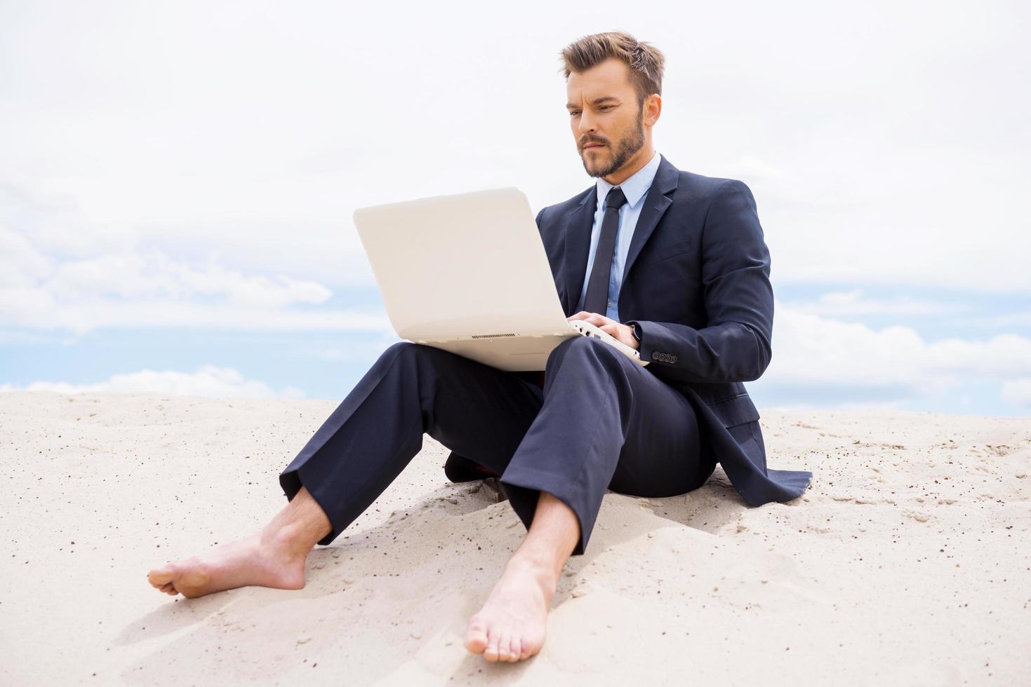 Businessman in desert. Confident young man in formalwear working on laptop while sitting on sand in desert photo
