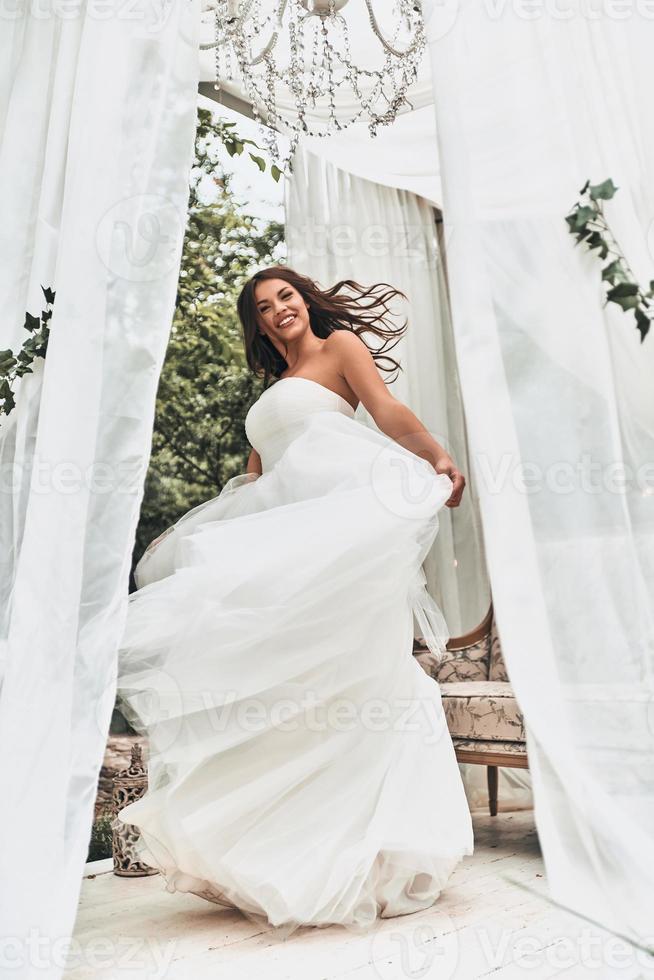 novia bailando. longitud completa de una joven atractiva con vestido de novia sonriendo y mirando a la cámara mientras gira al aire libre foto