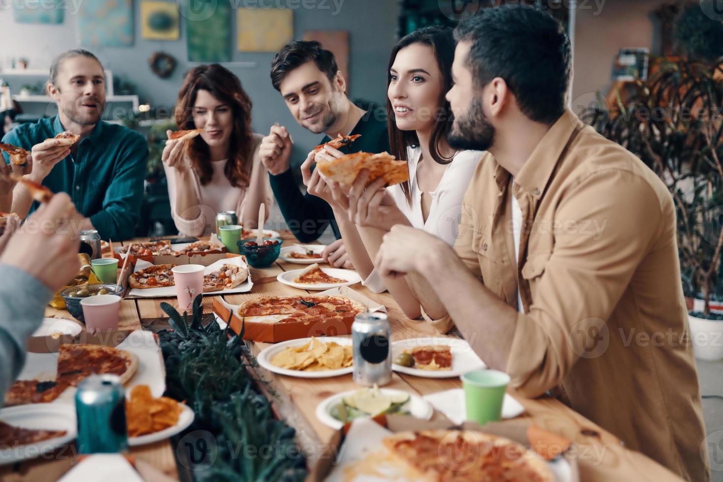 So tasty Group of young people in casual wear eating pizza and smiling while having a dinner party indoors photo